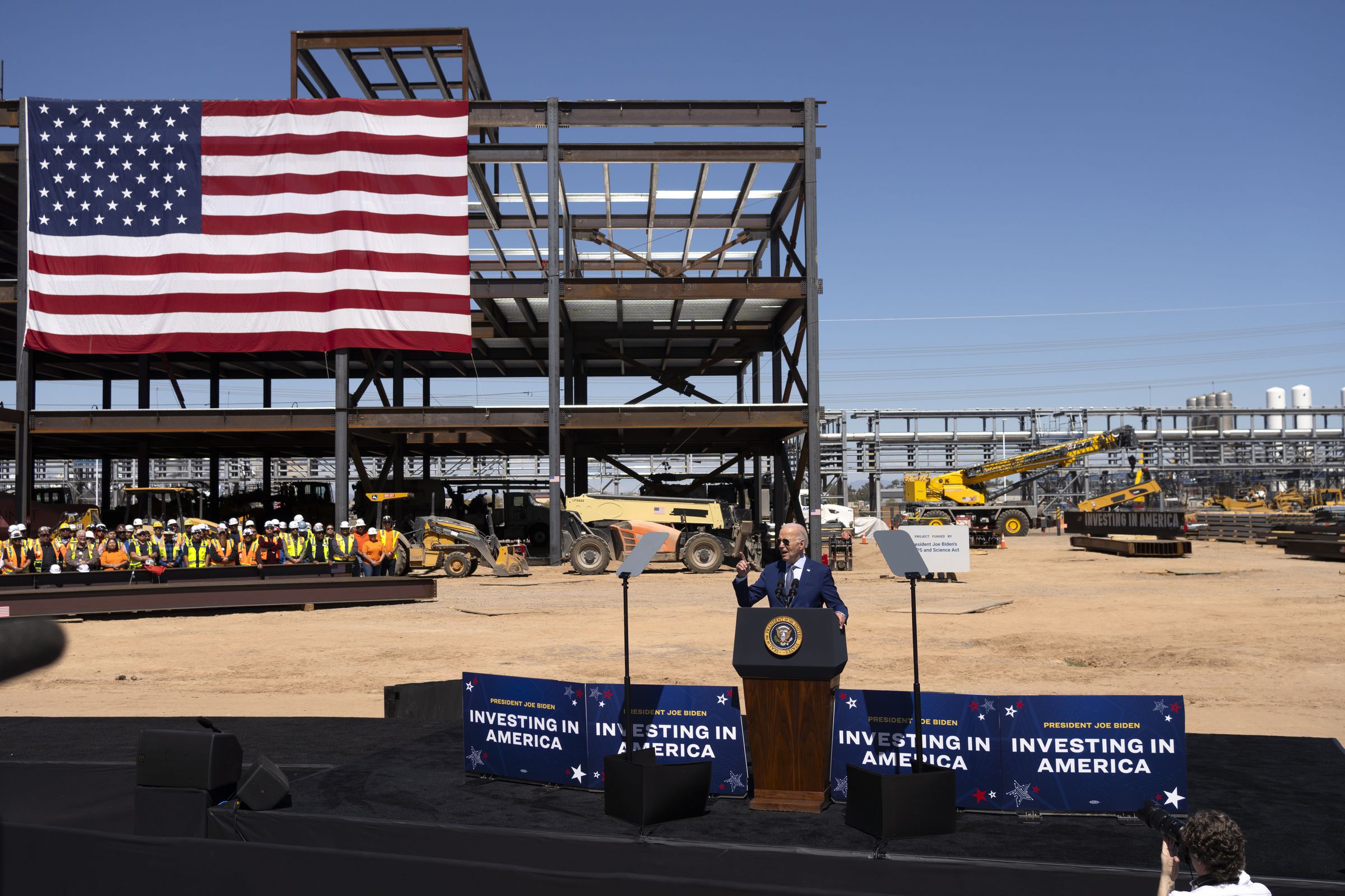 Joe Biden speaks at a podium in the middle of a construction site. Workers in bright vests and hard hats can be seen standing behind him, with a large American flag hung above them. Construction equipment is in the background.