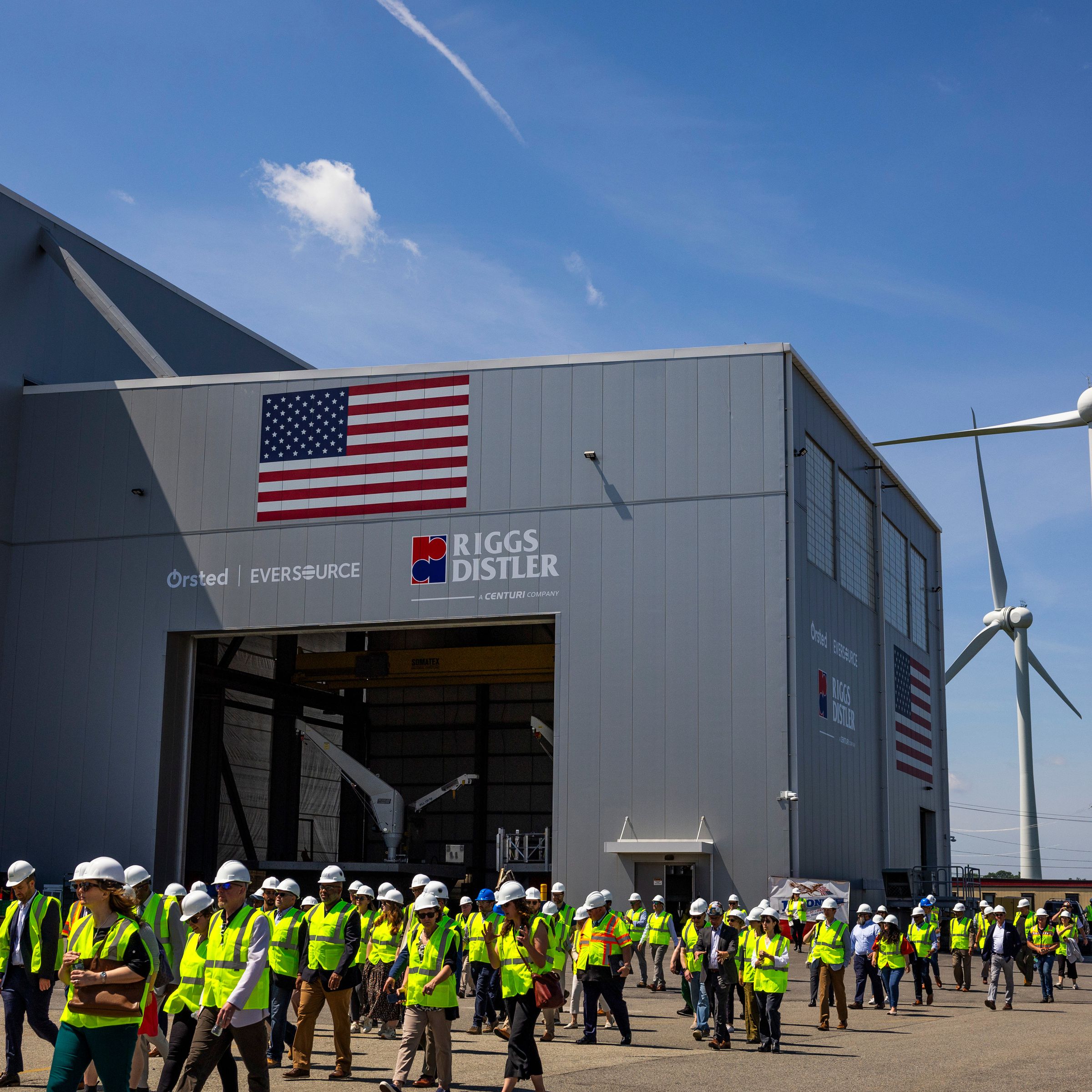 A line of people wearing construction vests and hard hats walk past a large industrial building with tall wind turbines seen behind them.