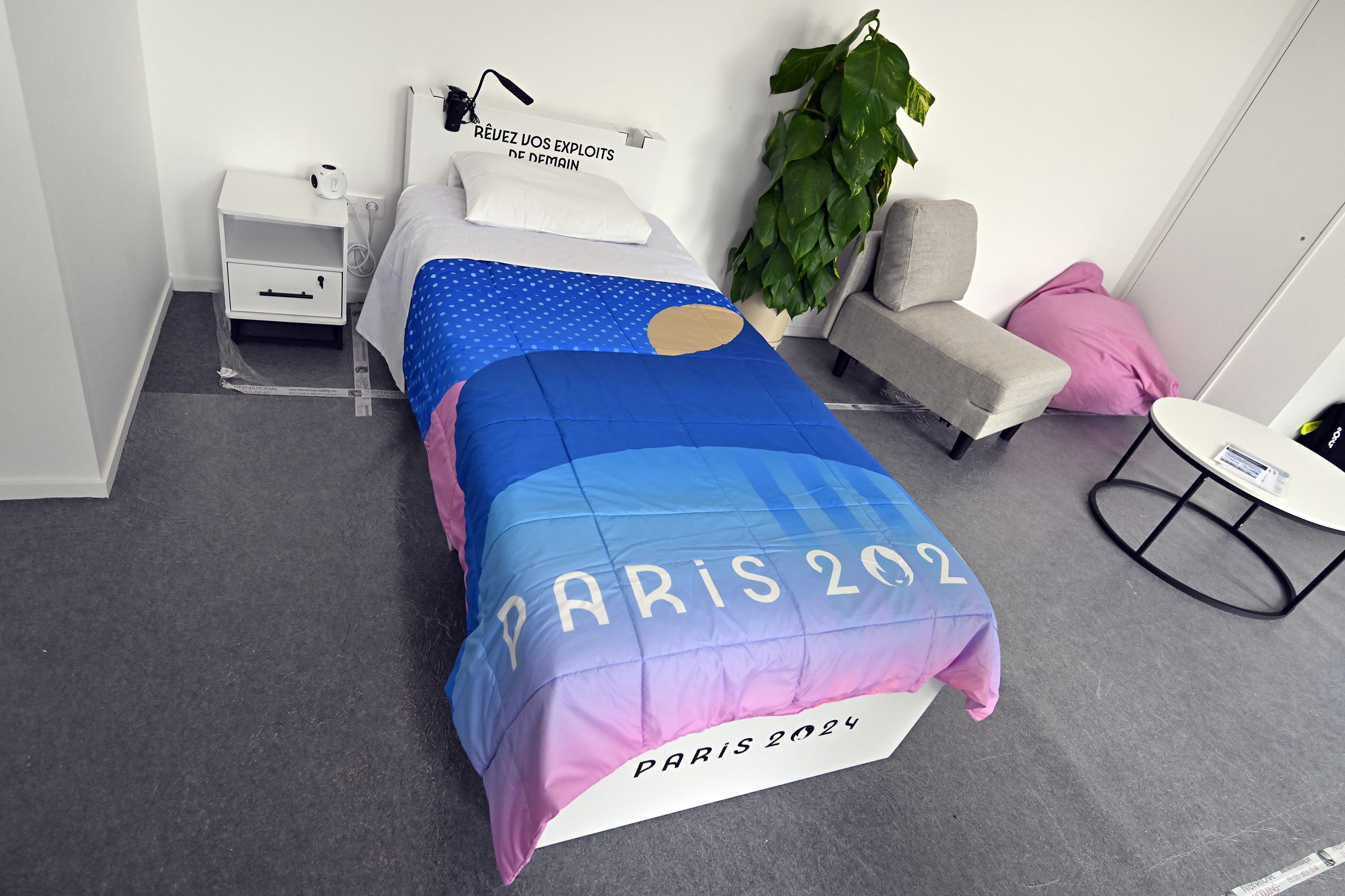 A room with a twin-sized bed covered with a blanket that says Paris 2024. There’s a nightstand on one side of the bed and on the other side a tall plant, a small lounge chair, and coffee table can be seen.