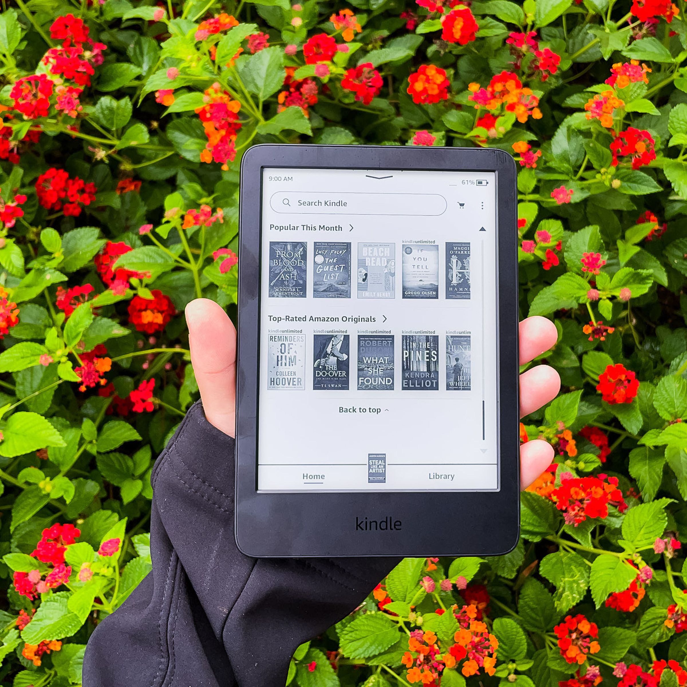 A hand holding the 2022 Kindle in front of red flowers.