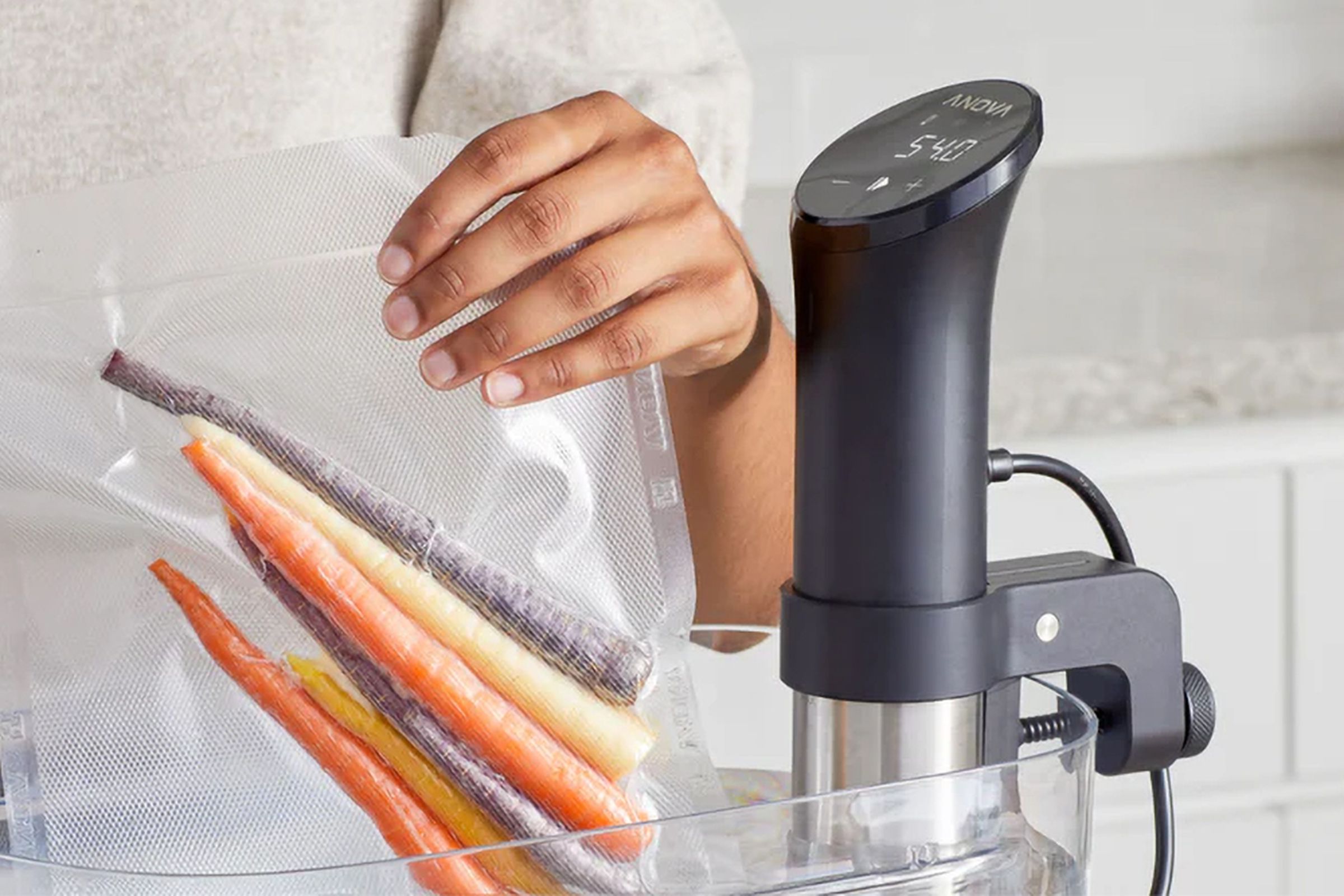 A person immersing a sealed bag of vegetables alongside an Anova sous vide machine.