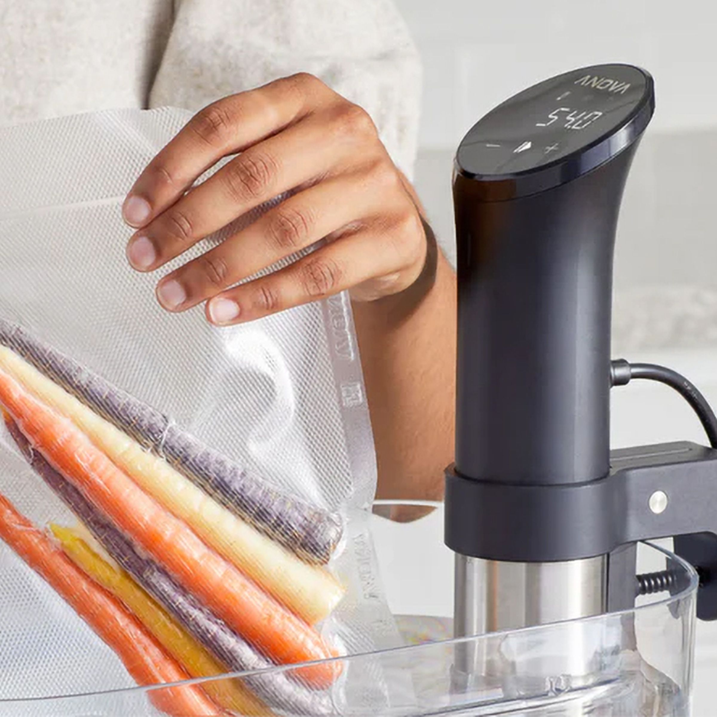 A person immersing a sealed bag of vegetables alongside an Anova sous vide machine.