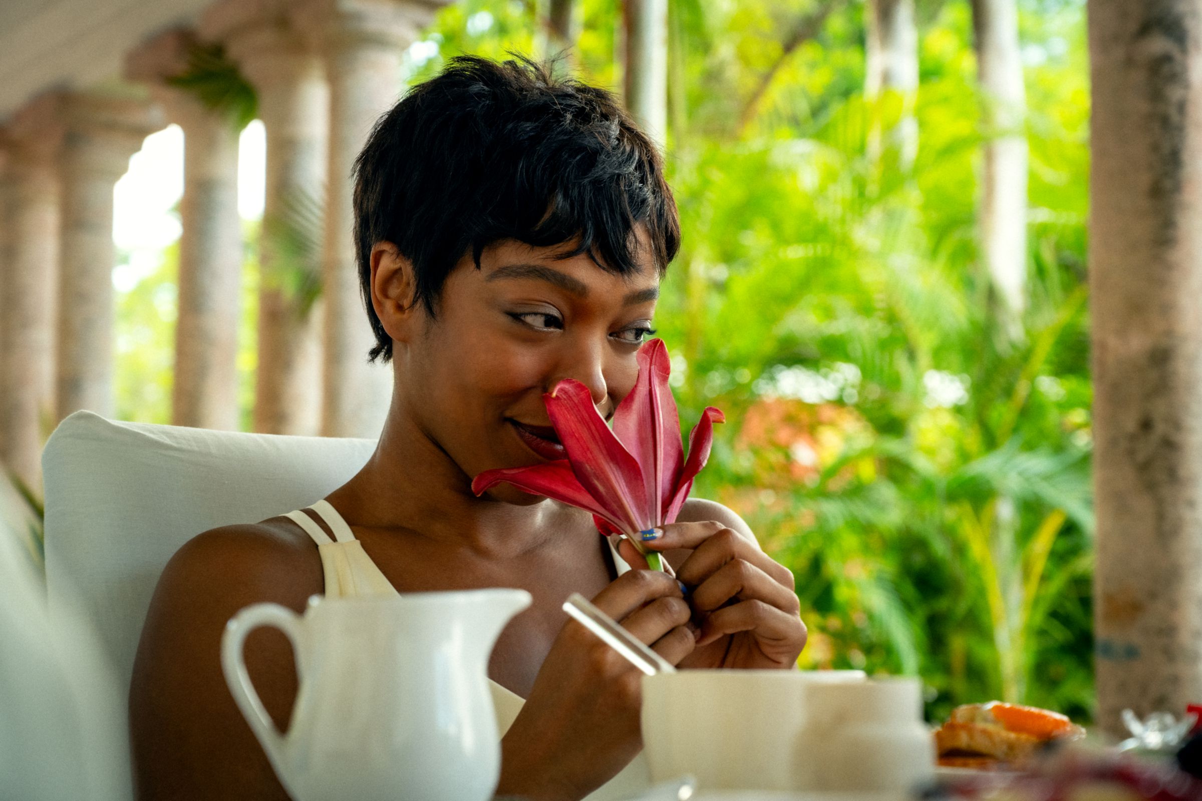 A woman sitting at a table sniffing a red flower.