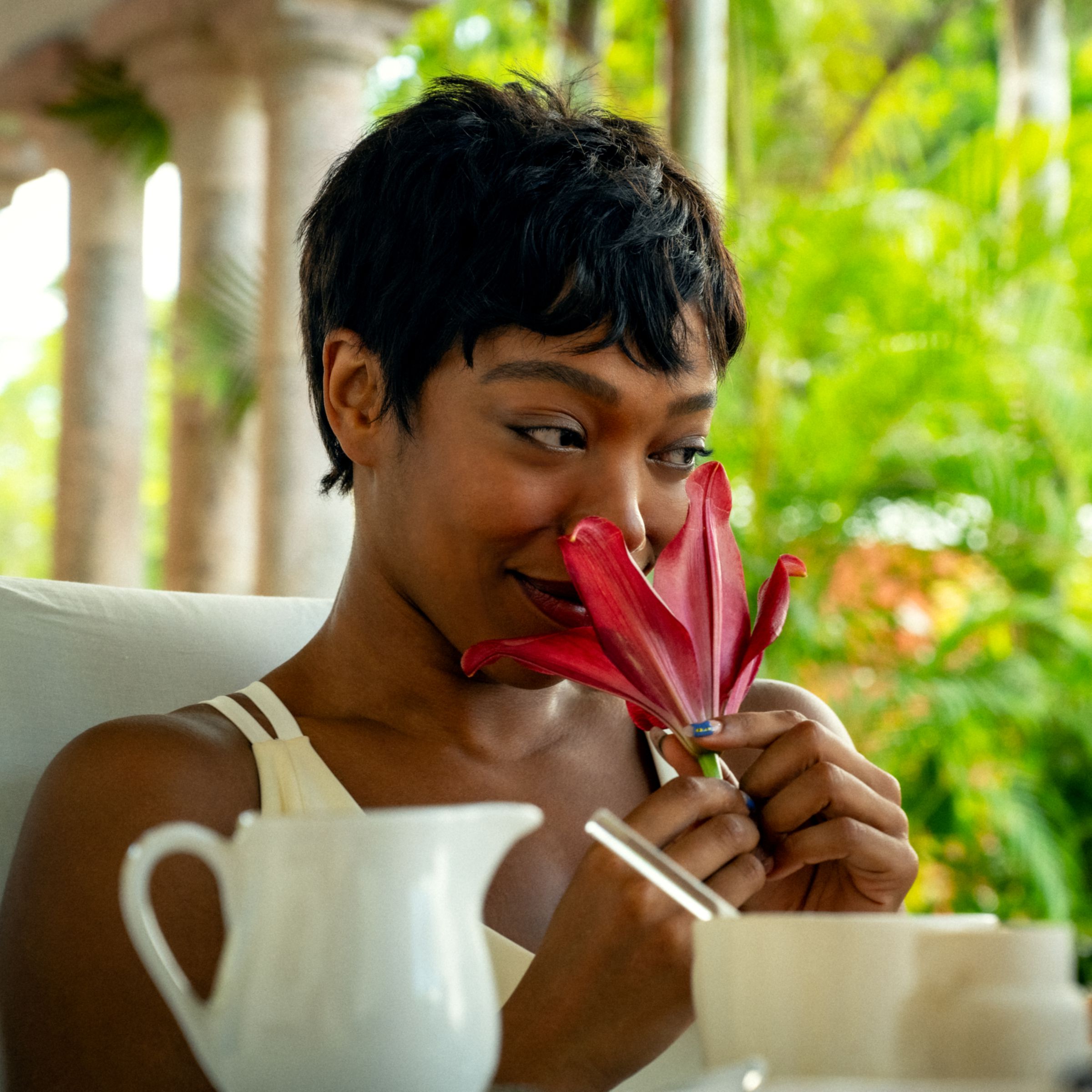 A woman sitting at a table sniffing a red flower.