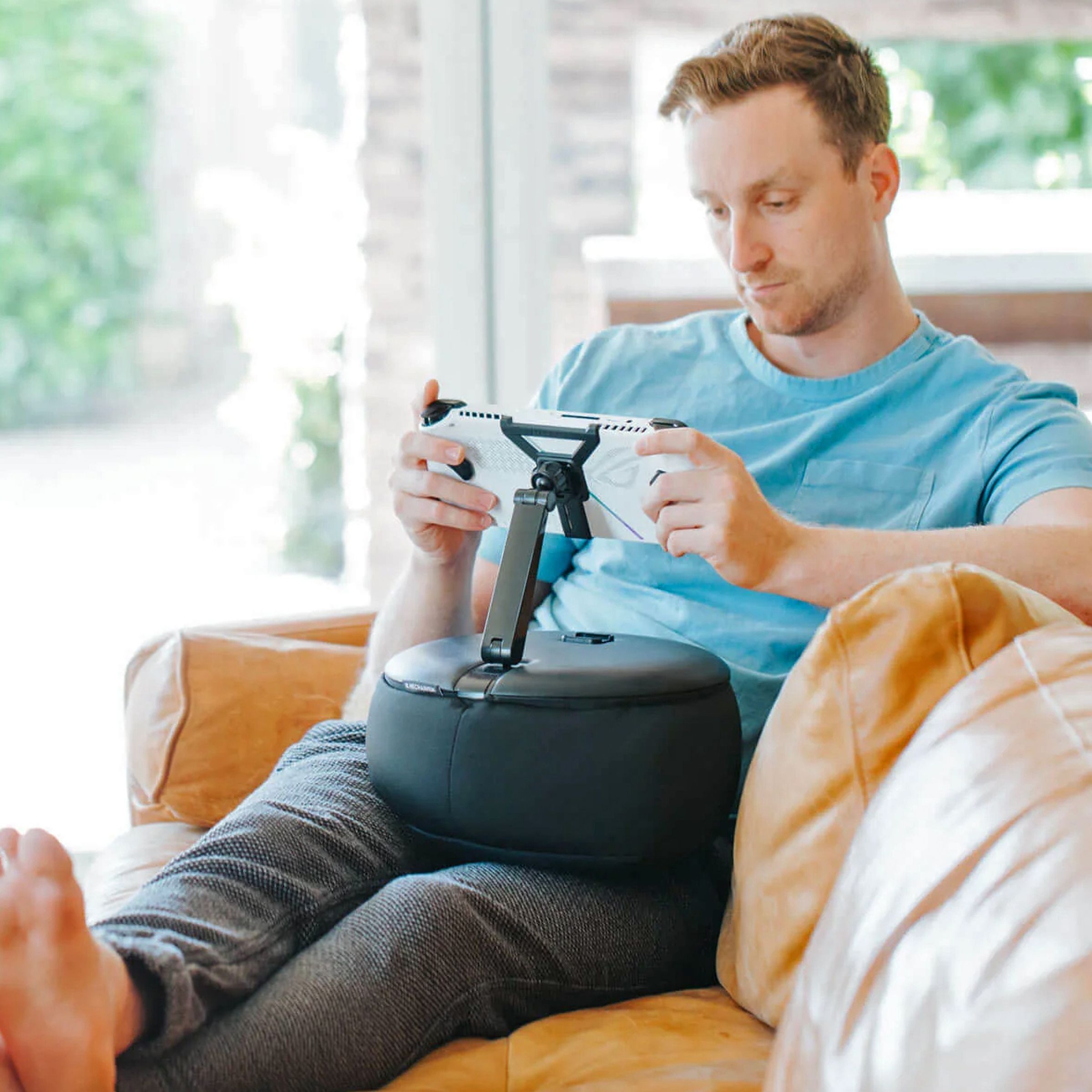 A gamer plays a handheld console on a sofa attached to the Gaming Pillow on their lap.