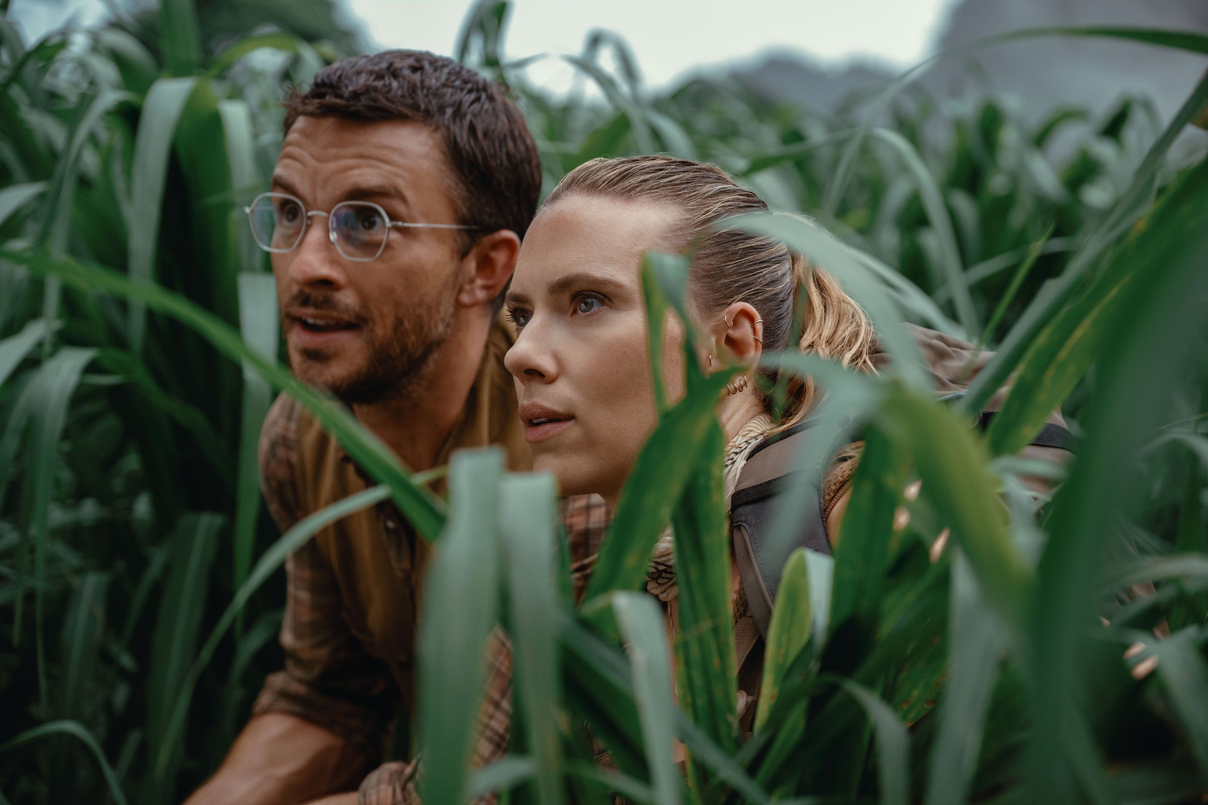 Production still from Jurassic World Rebirth featuring Jonathan Bailey and Scarlett Johansson crouching in tall grass.