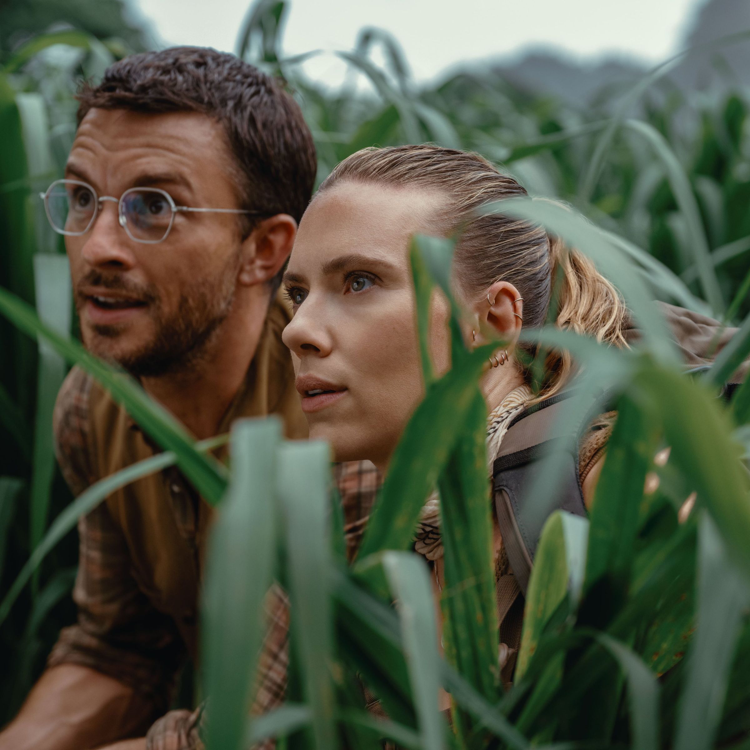 Production still from Jurassic World Rebirth featuring Jonathan Bailey and Scarlett Johansson crouching in tall grass.