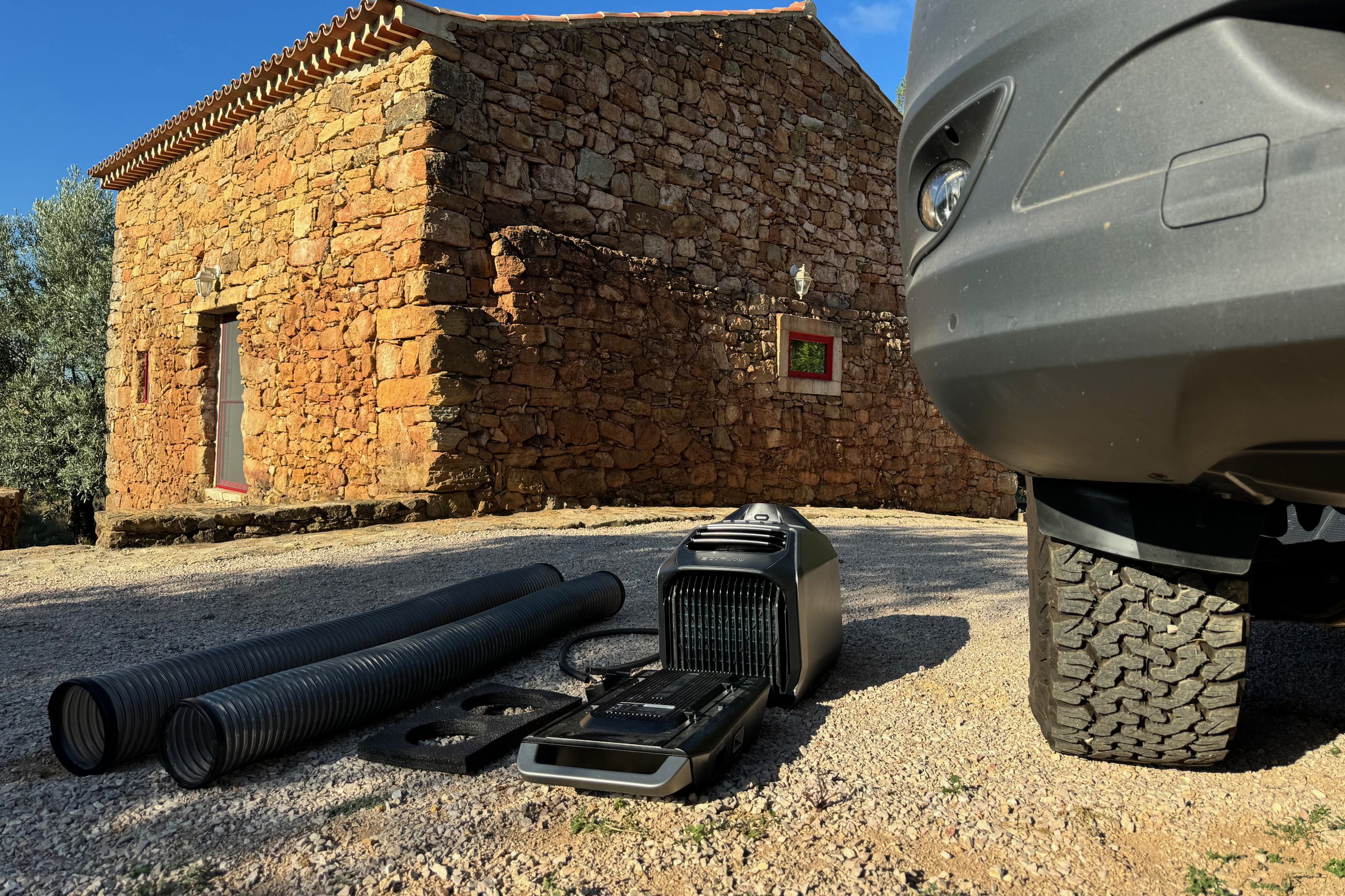 The Wave 2, its ducting, window vent board, drain and power cable sitting on gravel next to a large off-road wheel and an old stone farmhouse in the background.