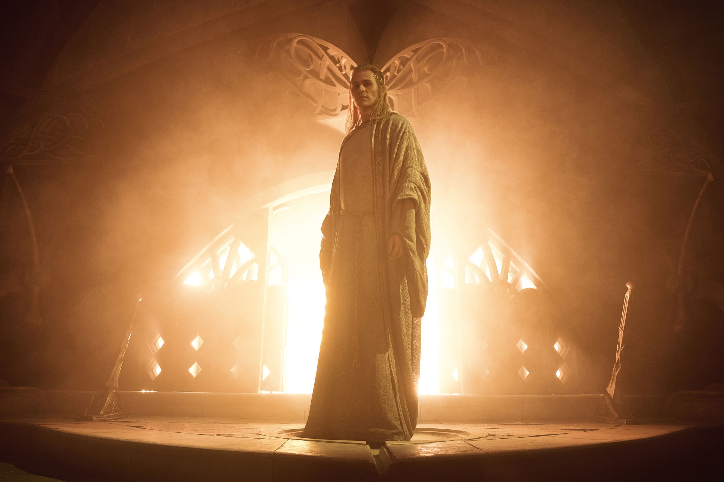 A man in a flowing white robe standing on a dais in front of a furnace from which flames are roaring and light is pouring.