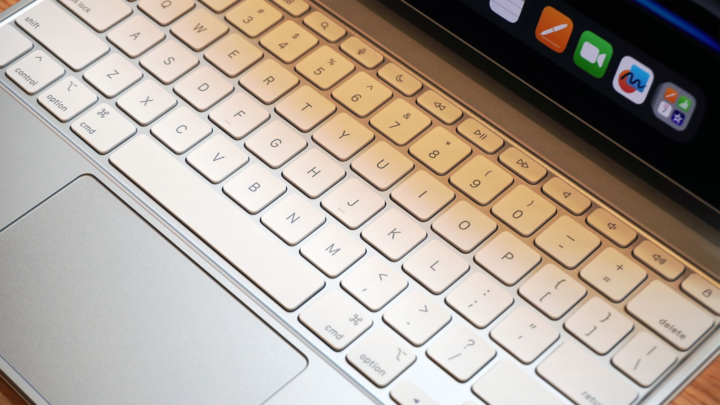 Close-up of a keyboard with a silver-colored top deck, white keys, and a large trackpad. It has a function row and is connected to an iPad Pro.